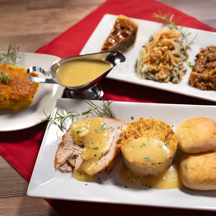 Thanksgiving dinner plate displaying 3 sides, pecan pie, and main course of turkey and gray with stuffing and rolls displayed on white plates, gravy in a gravy boat on top of a red napkin.