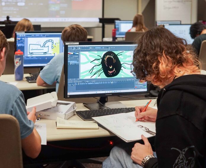 A female student drawing in a notebook while looking at a computer screen with an image in a classroom full of students.