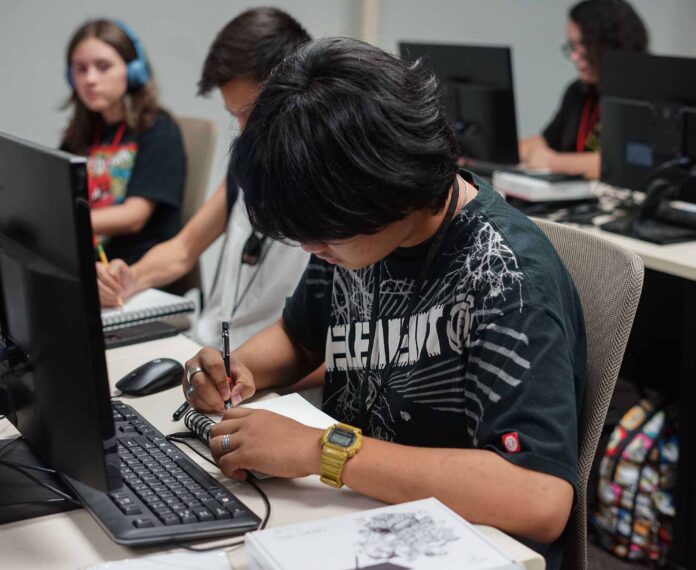 Male student drawing in his notebook sitting in front of a computer with other students in the background in a computer lab classroom.