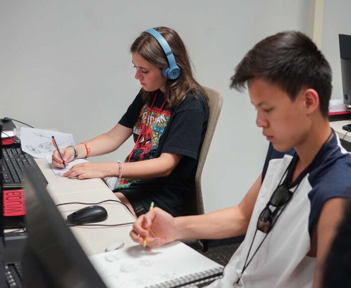 two students drawing on notebooks sitting in front of computers in a lab