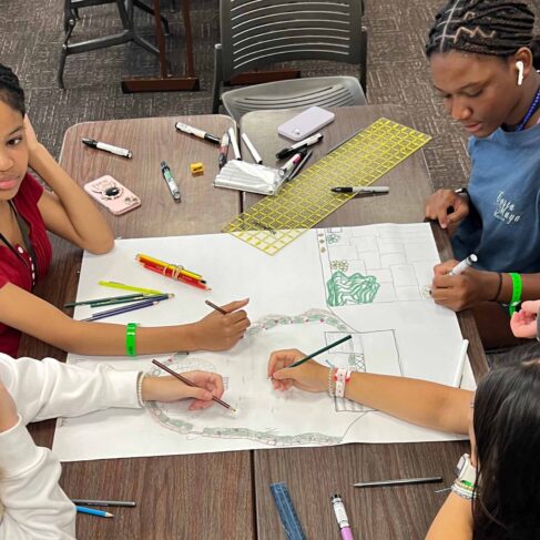 five students designing a building structure on a piece of paper on a table in a classroom