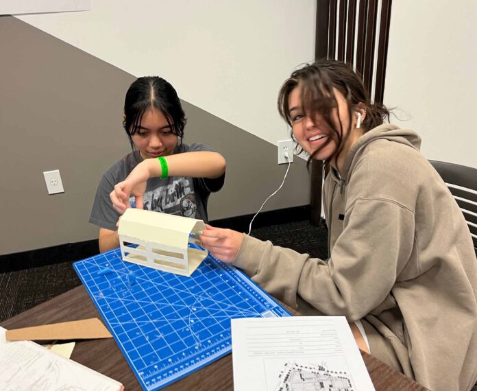 two students building a structure on a table by following a diagram in a classroom