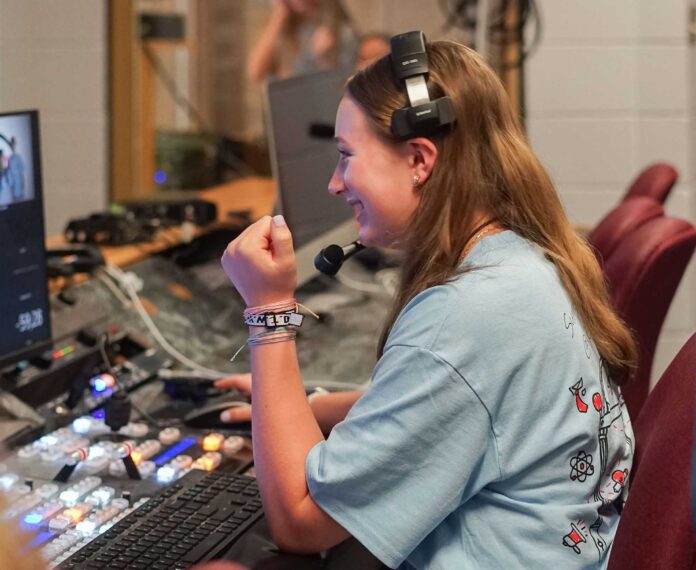 A student sitting at a control panel wearing headphones is happy looking at a screen.