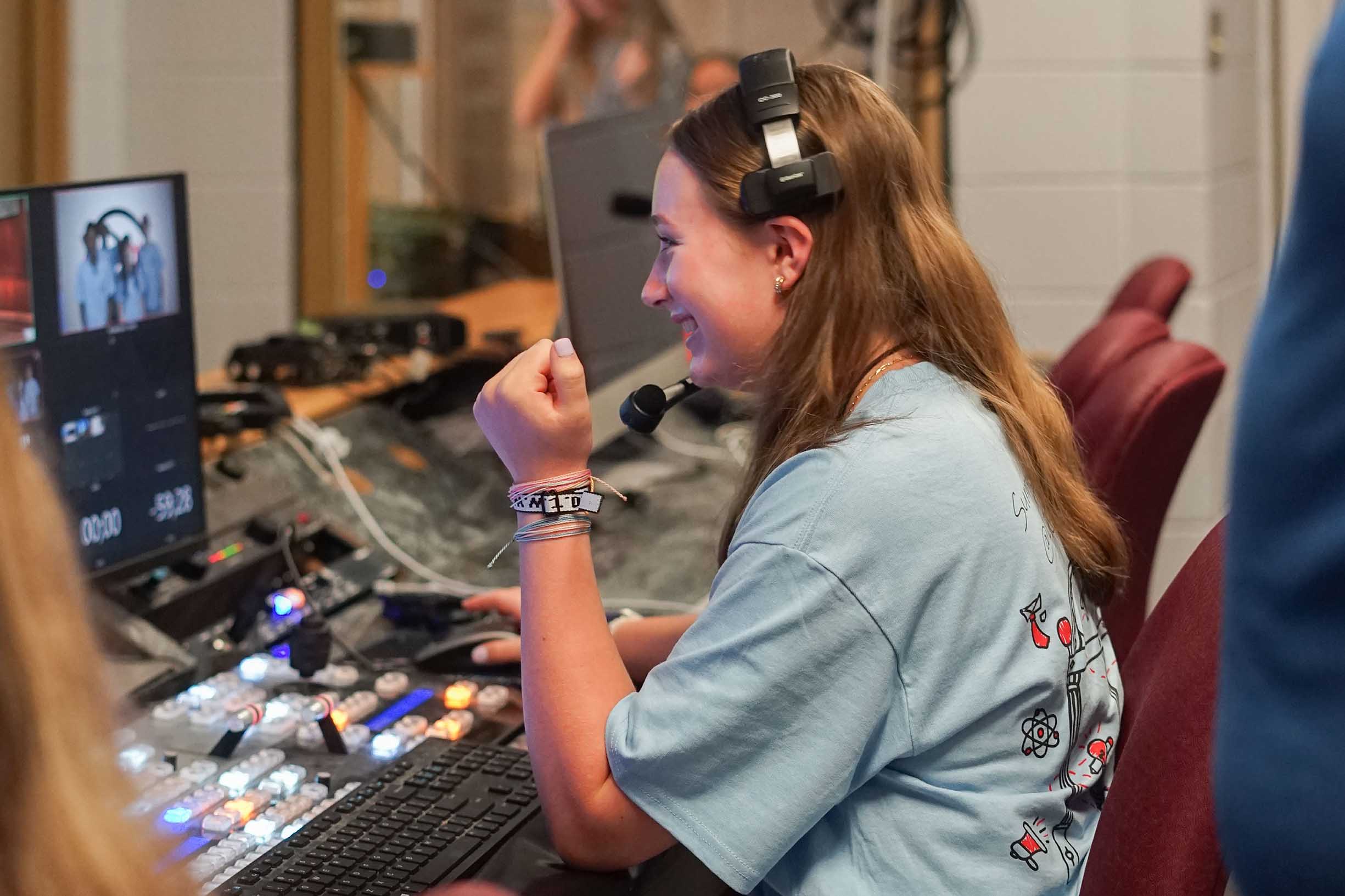 A student sitting at a control panel wearing headphones is happy looking at a screen.