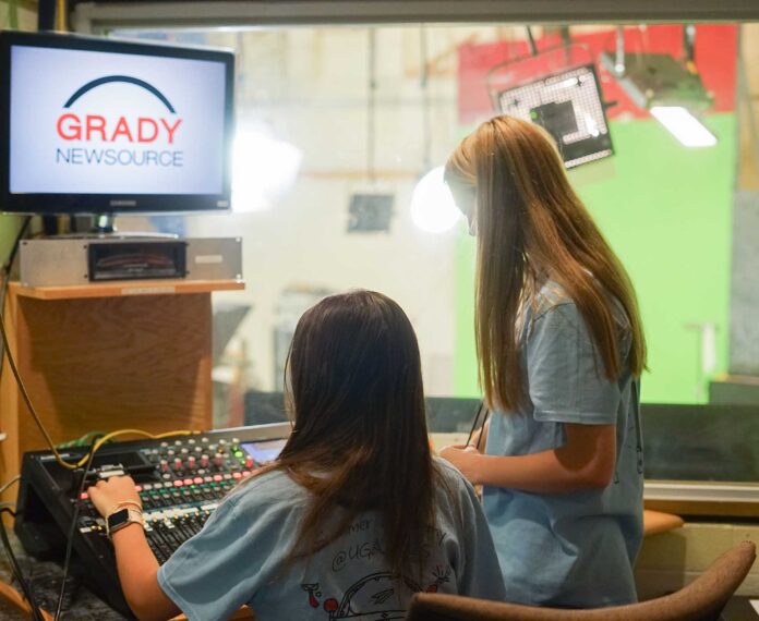 Two students in a studio looking at a control panel with a computer screen with the "Grady Newsource" logo.