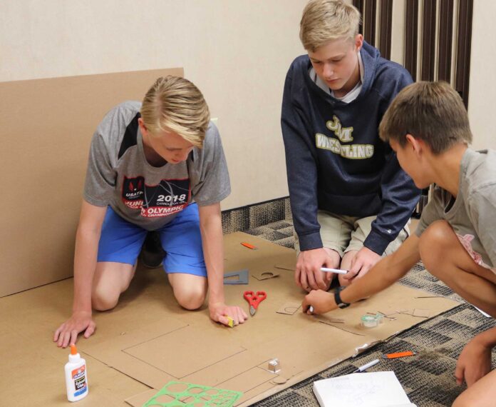Three male students cutting cardboard on the floor in a classroom.