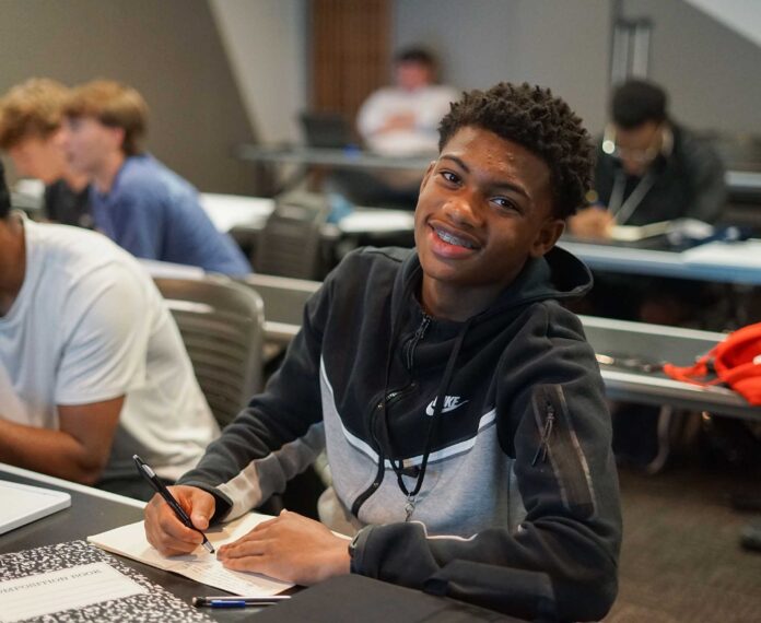 A male student sitting in a classroom writing in a notebook.