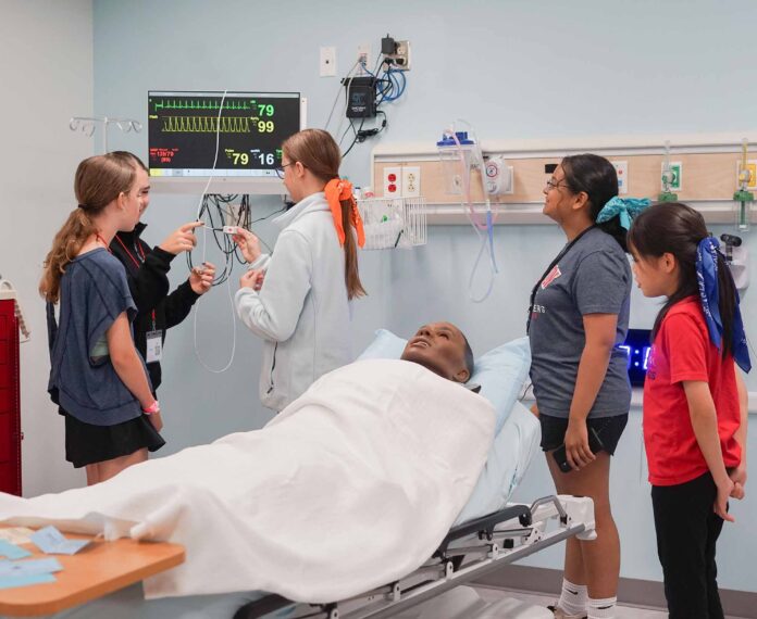 five students in a medical classroom with a practice dummy laying on the medical bed