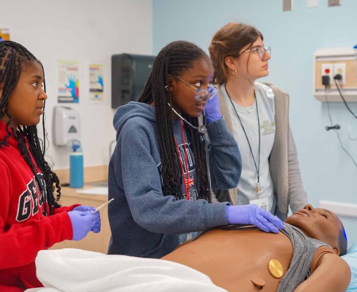 three students in a medical classroom with a practice dummy laying on the medical bed