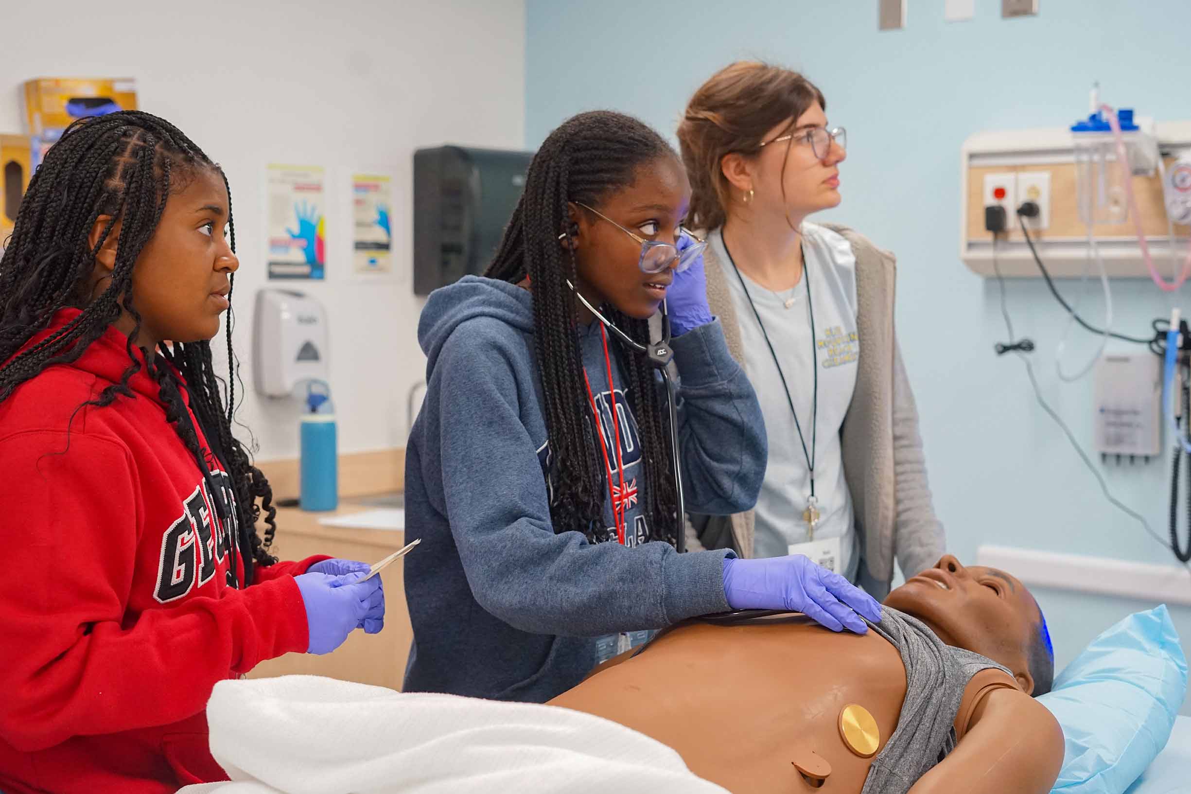 three students in a medical classroom with a practice dummy laying on the medical bed