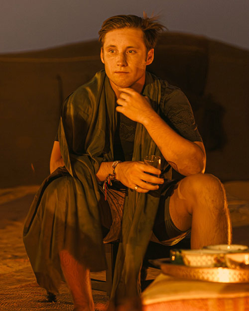 Sam sitting outside in front of a hilly area wearing a dark shirt and shorts with a blanket draped over his shoulders.