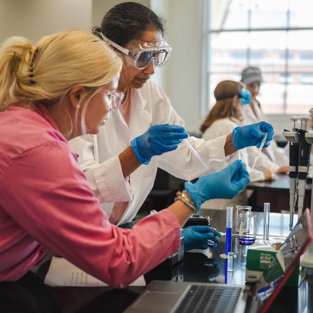 two students working on an experiment using pipettes and beakers with blue fluid in a lab on UGA campus
