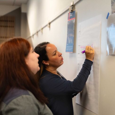 Two women writing on a large sheet of paper that taped to a white board.