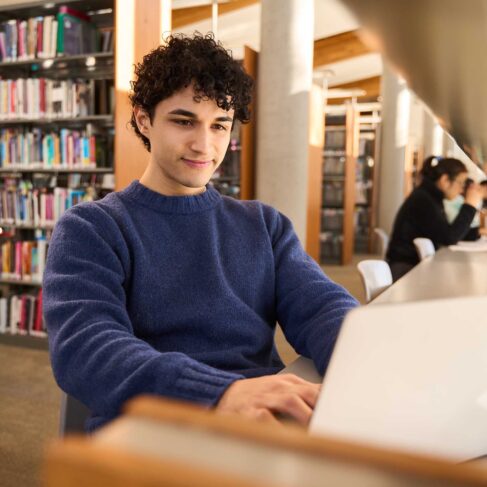 Man working on laptop in a library with other students studying in background