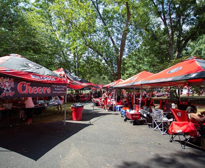 A tailgating scene under trees, with rows of red and black Georgia Bulldogs canopy tents lining both sides of a paved path. People in red clothing sit in folding camp chairs and stand near tables of food, with coolers, wagons, and a red trash bin nearby.