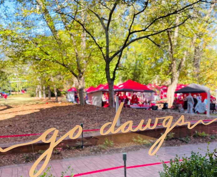 "Go dawgs" in gold script on a window, looking out to red tailgate tents under trees on a sunny campus.